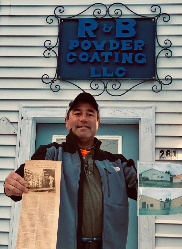 Rob Hewes, founder of R&B Powder Coating, stands in front of his business holding the original newspaper clipping announcing the construction of his new building and loan assistance from REDC in 2004.